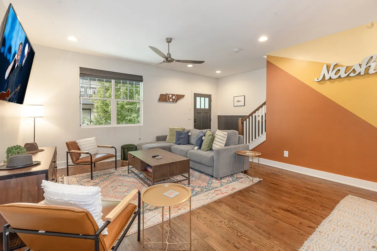 The image showcases a stylish living room with a sofa adorned with colorful cushions, a decorative rug beneath a modern coffee table, and a large TV mounted on the wall. The space feels bright, welcoming, and perfect for relaxing or gathering with friends and family.