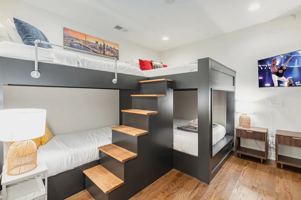 A playful and inviting bunk room with two sets of bunk beds, each equipped with lamps, a TV mounted on the wall, and a built-in staircase for easy access. The room exudes warmth and is designed for both relaxation and fun.