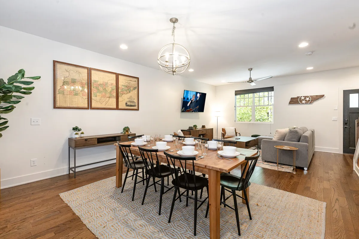 This image captures a welcoming living and dining area, featuring a dining table elegantly set for a meal, a comfortable sofa in the corner, decorative wall art, and a television mounted on the wall. The space is flooded with natural light, contributing to an inviting and sociable atmosphere.
