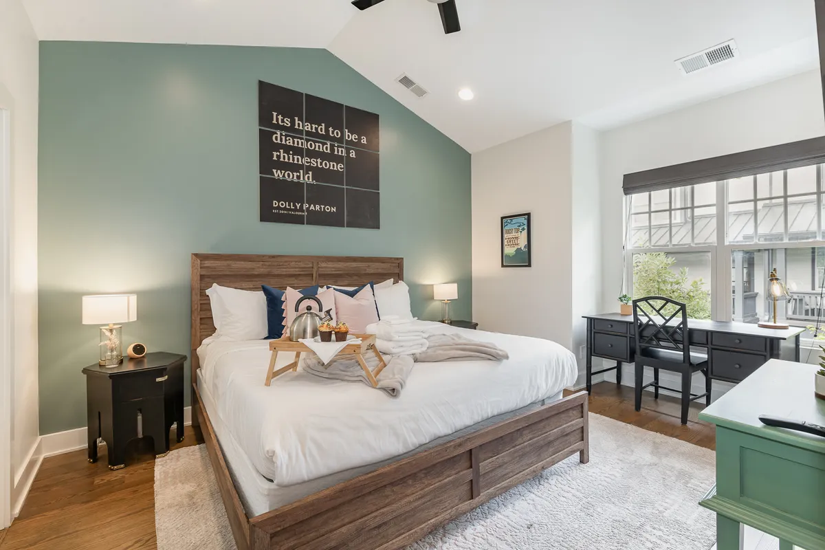 A sunlit bedroom featuring a comfortable bed with decorative pillows, a motivational Dolly Parton quote art piece above the headboard, a desk with a lamp and chair by the window, and soft ambient lighting. The design is welcoming and inspirational, ideal for winding down or energizing mornings.