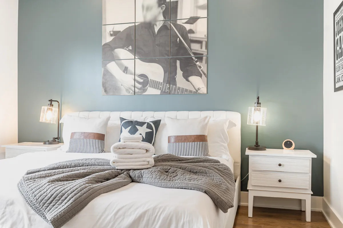 A bright, serene bedroom featuring a large bed with white linens, a nightstand with a lamp and clock, and framed artwork of a musician. The ambiance is calm and welcoming, ideal for unwinding after a busy day.