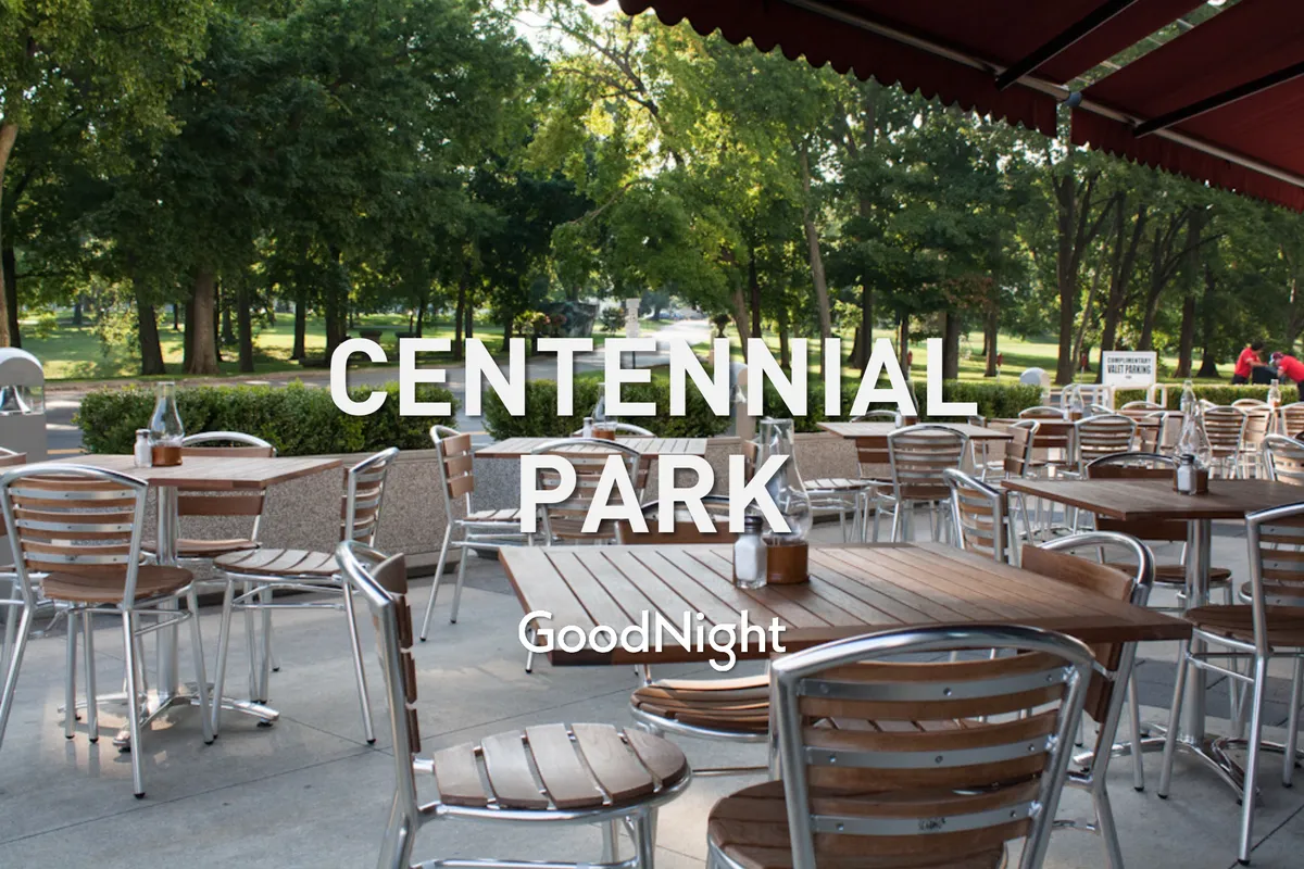 This exterior image showcases a relaxed outdoor seating area with wooden tables under a canopy awning, surrounded by green trees. The scene is casual and social, suggesting a space where guests can gather, dine, or unwind outdoors.