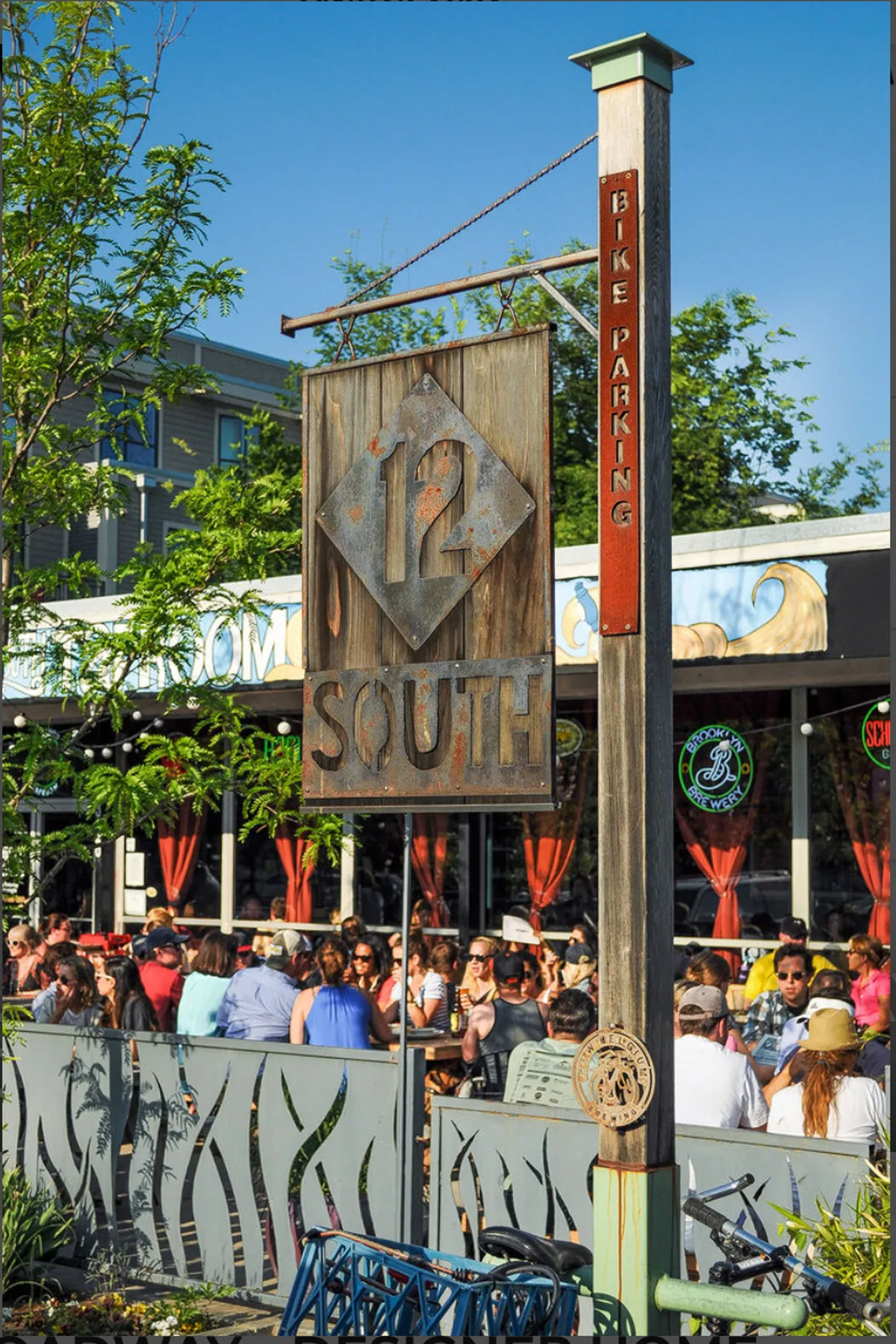 A vibrant streetscape from the 12 South district featuring the prominent 12 South sign, a bike parking sign, busy outdoor seating, and people dining and socializing. The scene is lively, with a blend of local shops, eateries, and a strong sense of neighborhood activity.