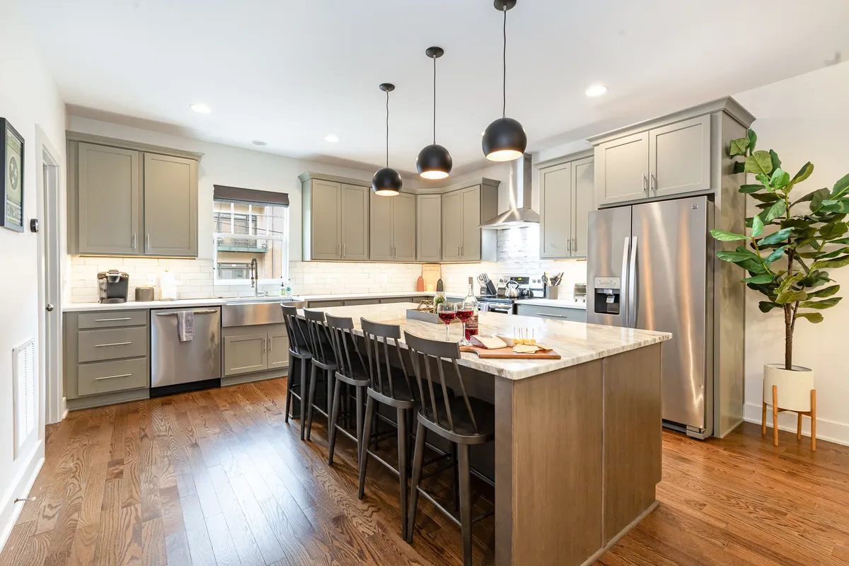 A large, open kitchen island is the centerpiece of this image, complemented by pendant lights, stainless steel appliances, and a touch of greenery. The space is clearly designed for cooking, entertaining, and casual dining.